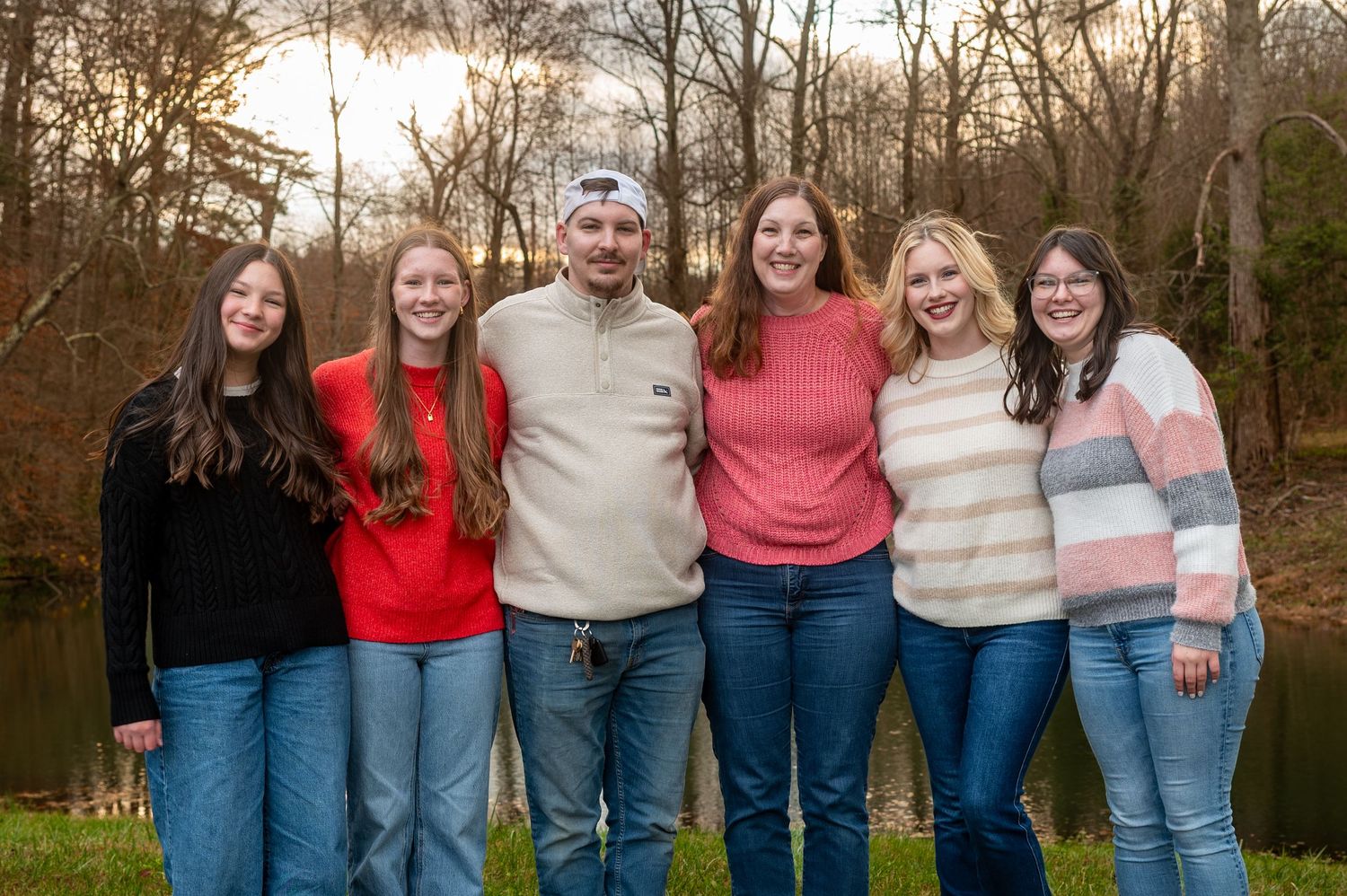 Family of six smiling outdoors near a pond with bare winter trees in the background.