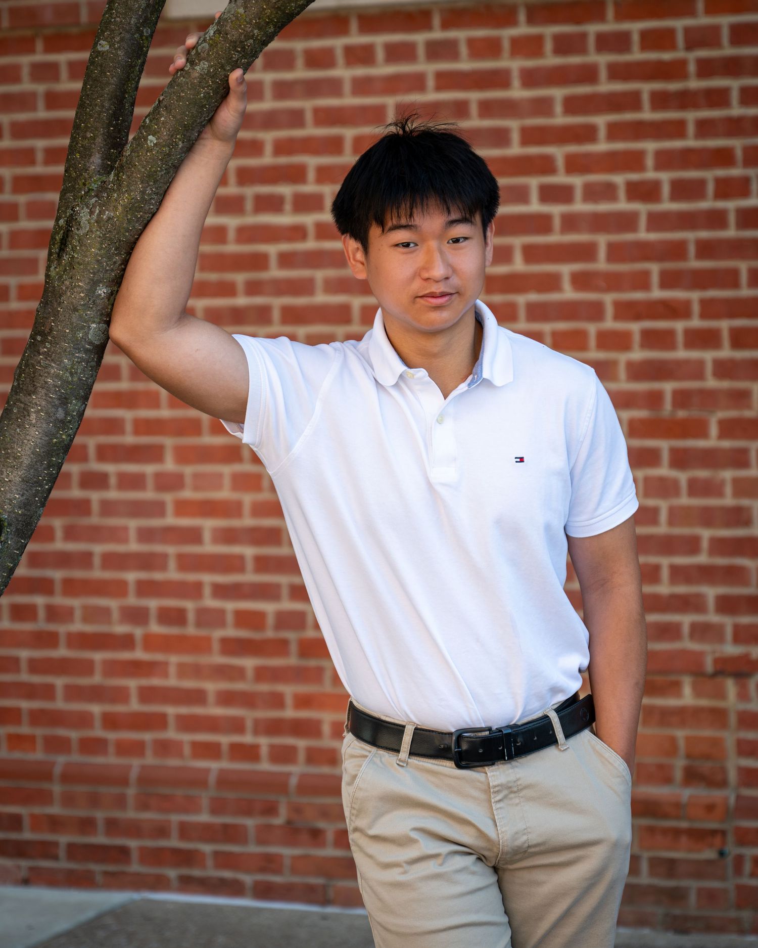 Young man in white polo shirt and khaki pants leaning against a tree in front of a brick wall.
