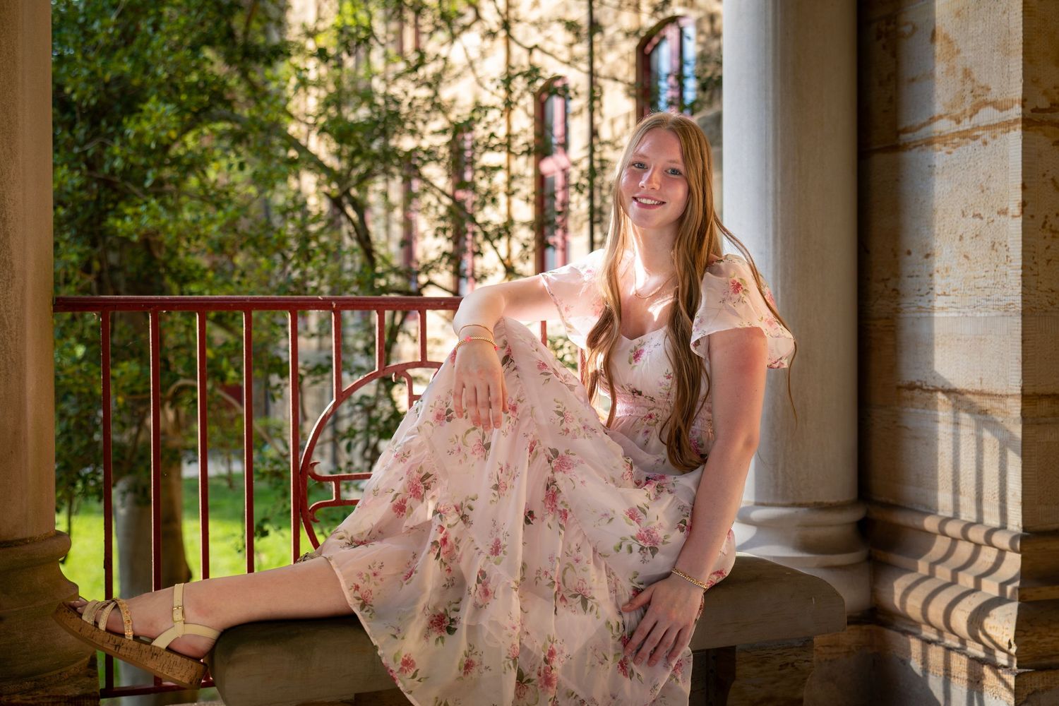 Young blonde woman in floral dress poses gracefully on ornate balcony with red railing and lush greenery.
