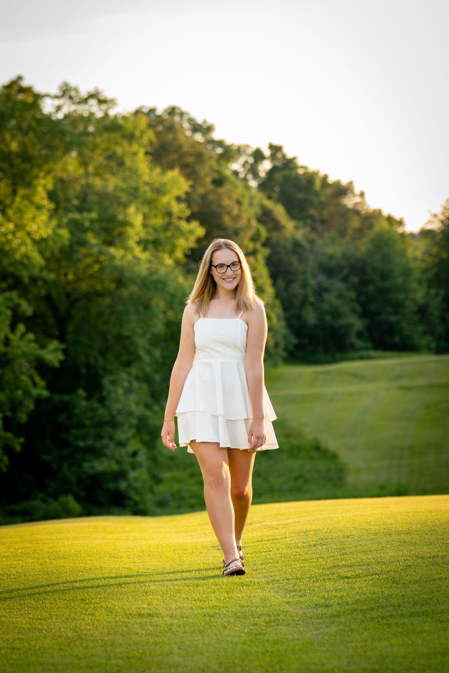 Young woman in white sundress and glasses walking on lush green grass with trees in background.