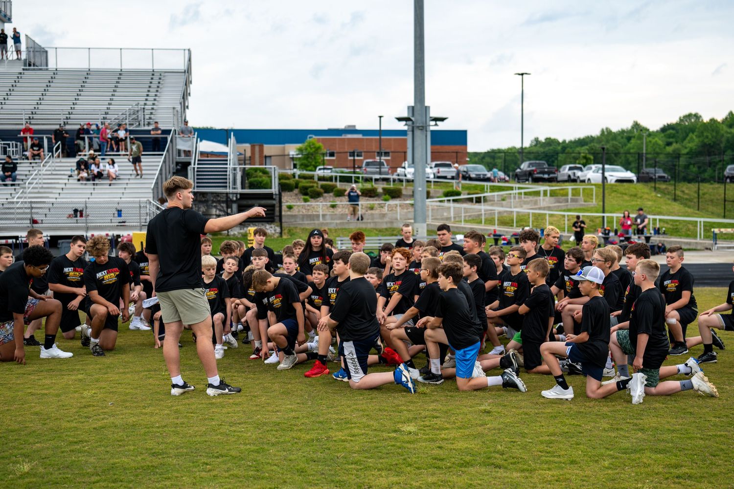 Large group of youth athletes in black shirts kneeling on a field during an outdoor sports camp drill.