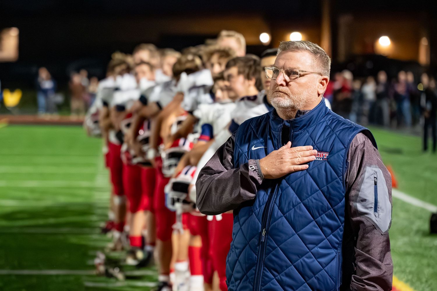 Coach stands with hand over heart during national anthem alongside football players lined up on sideline.