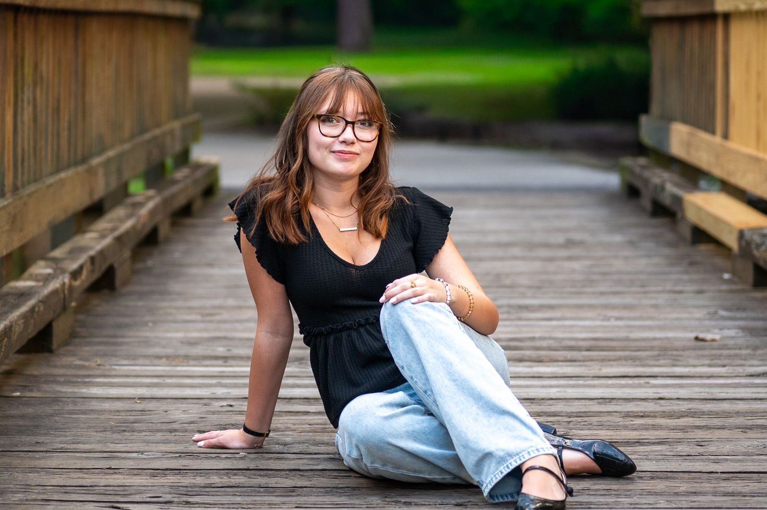 Young woman with glasses sitting on a wooden bridge in a park, wearing a black top and light blue jeans.