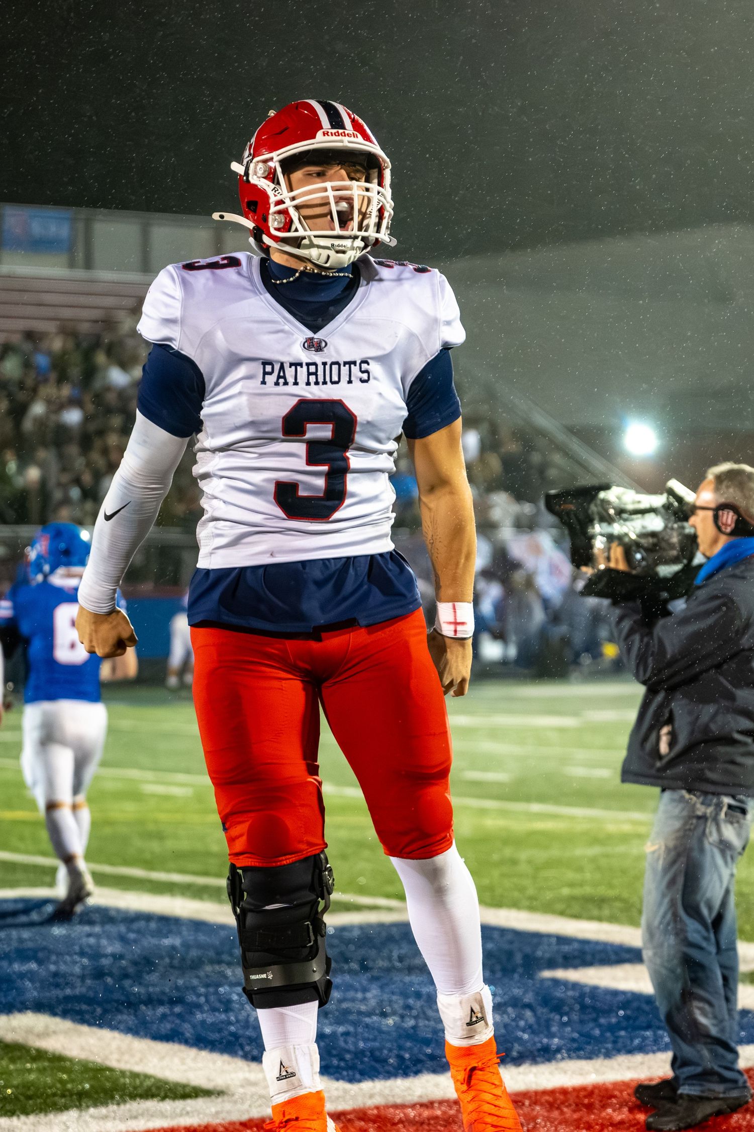 Football player wearing Patriots number 3 jersey and helmet celebrating on a rainy night game field.