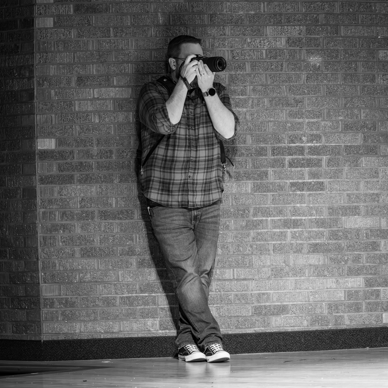 Black and white photo of Torrey Hanke, co owner of Hanke Studios, leaning against a brick wall holding a camera to his face.