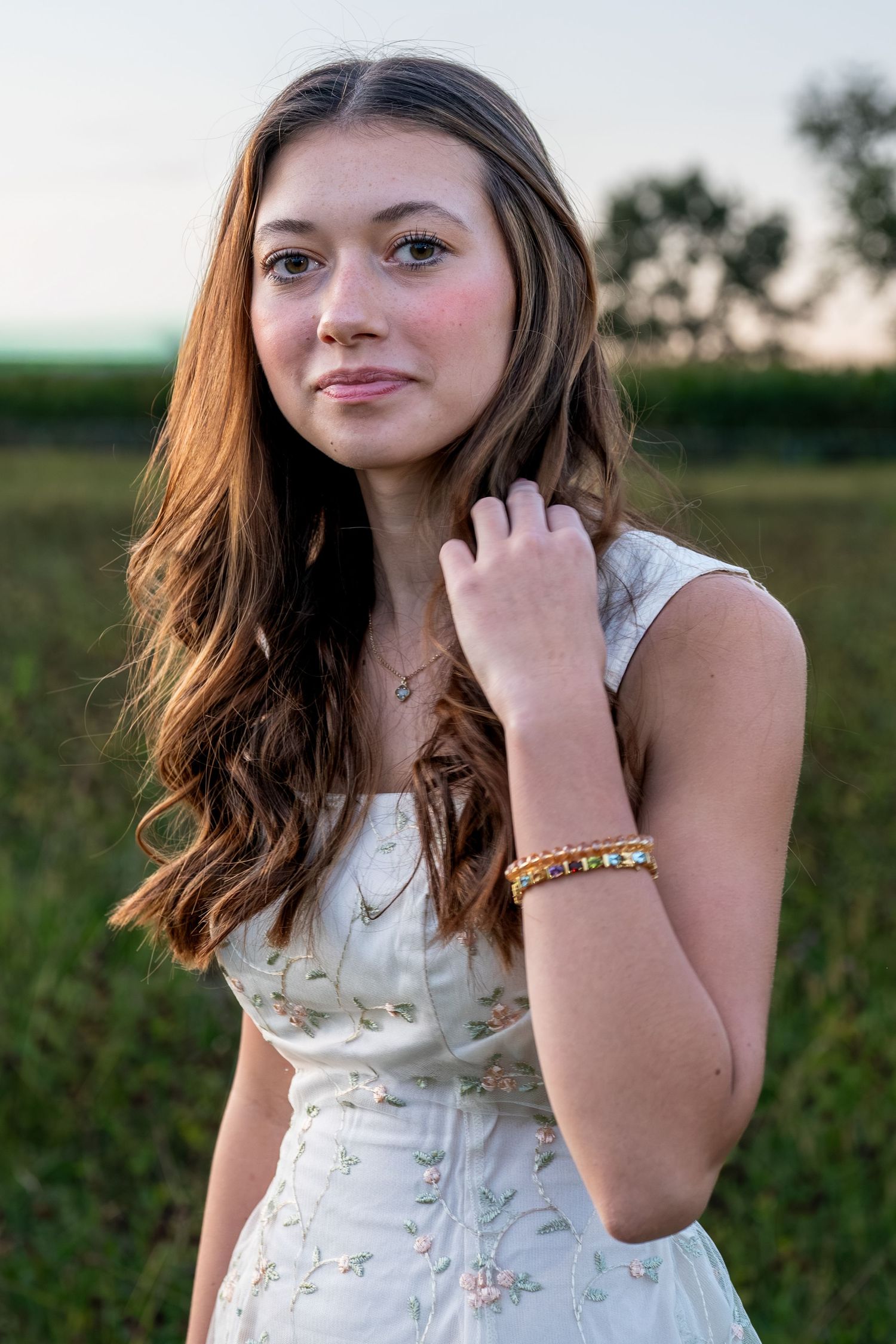 Young brunette woman in white floral dress holds her hair back in a golden hour field.