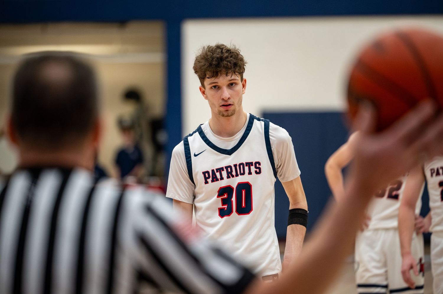 Patriots player #30 focuses during a high school basketball game with a referee in the foreground.