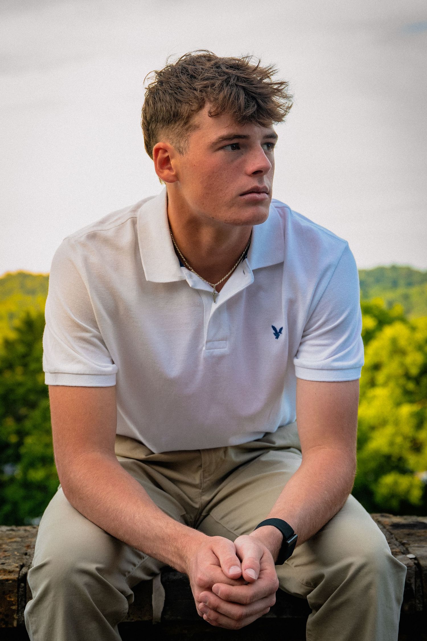 Young man in white polo shirt sitting outdoors with hands clasped, looking away thoughtfully with green trees behind him.