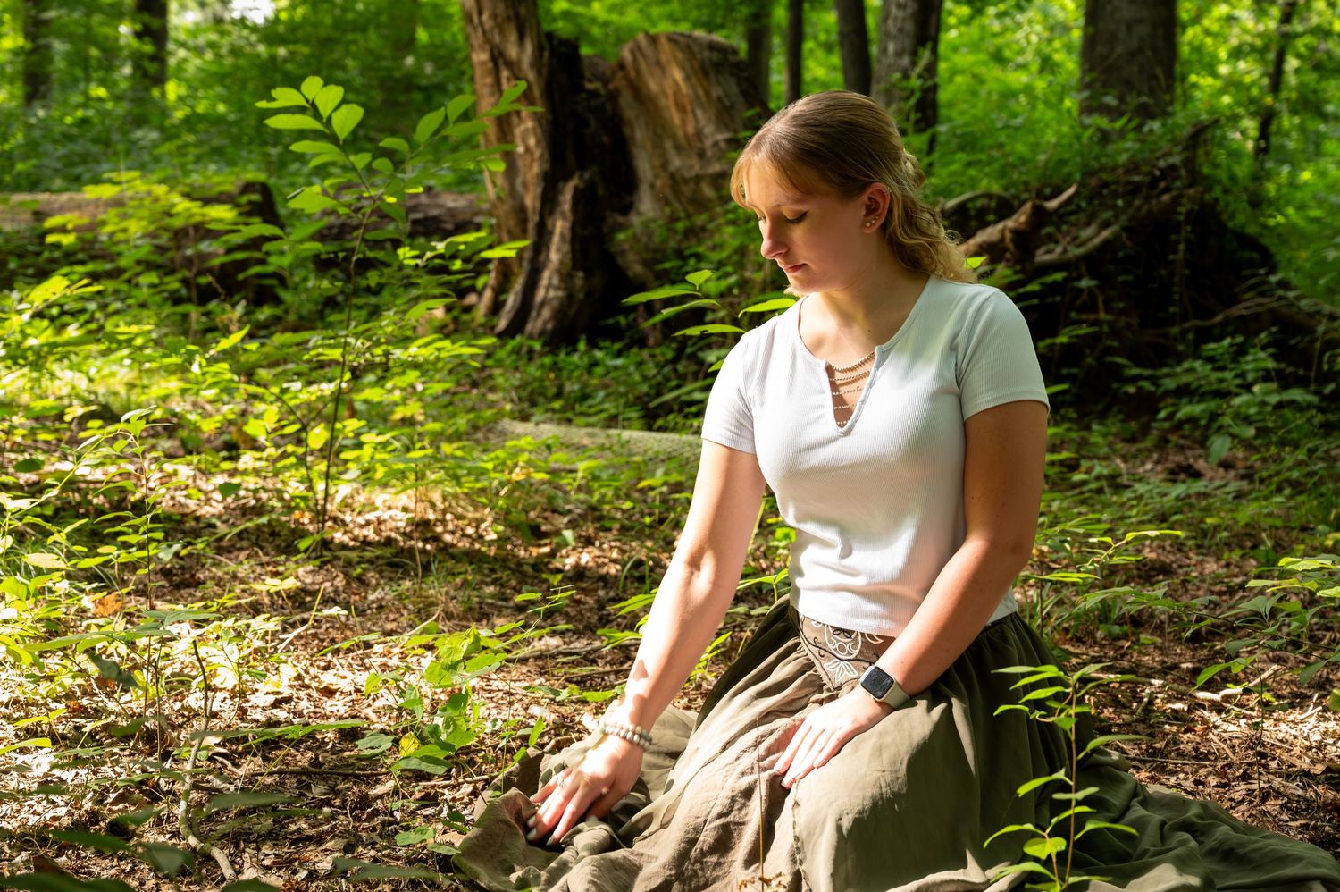 Young woman in white top and olive skirt kneeling peacefully in a sunlit forest surrounded by green foliage.