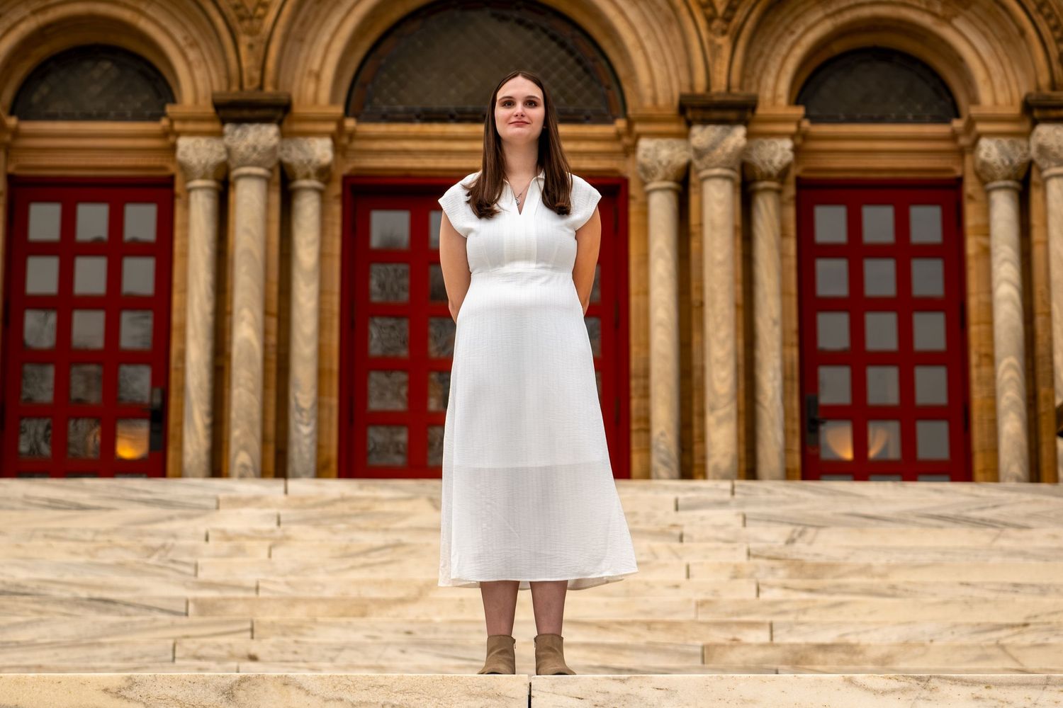 Woman in white midi dress standing on marble steps before ornate building with red doors and columns.