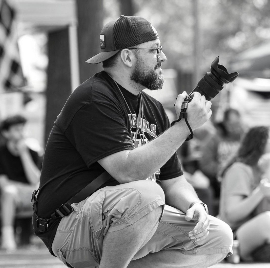Torrey Hanke, co owner of Hanke Studios, crouching outdoors with camera at event, captured in black and white side profile.