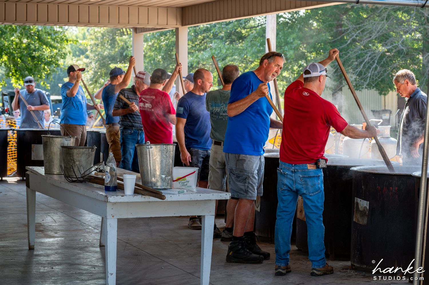 Men cooking large pots of food outdoors at a community event under a covered pavilion.
