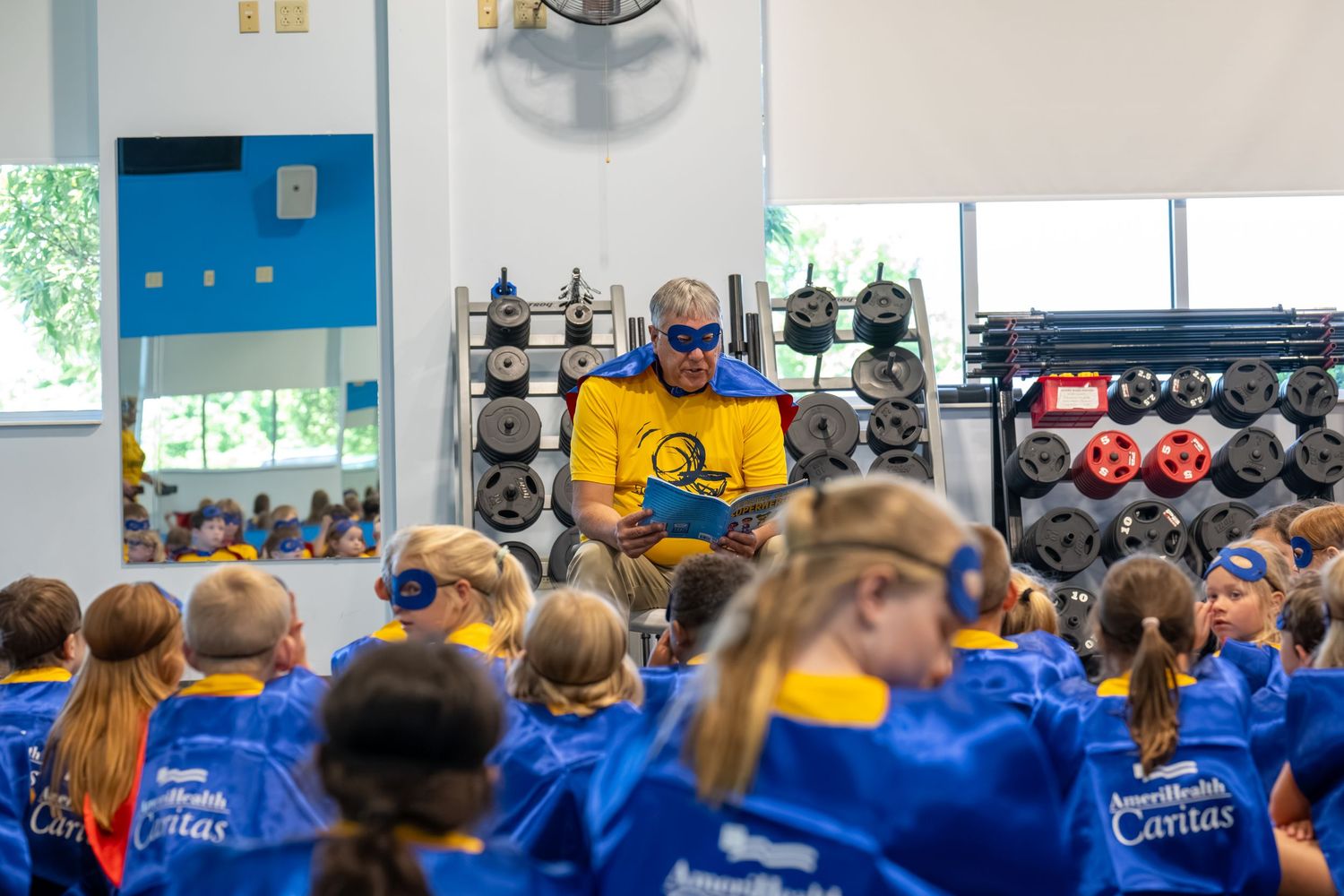 Superhero-costumed reader engages young students in blue and gold uniforms during a school reading event indoors.