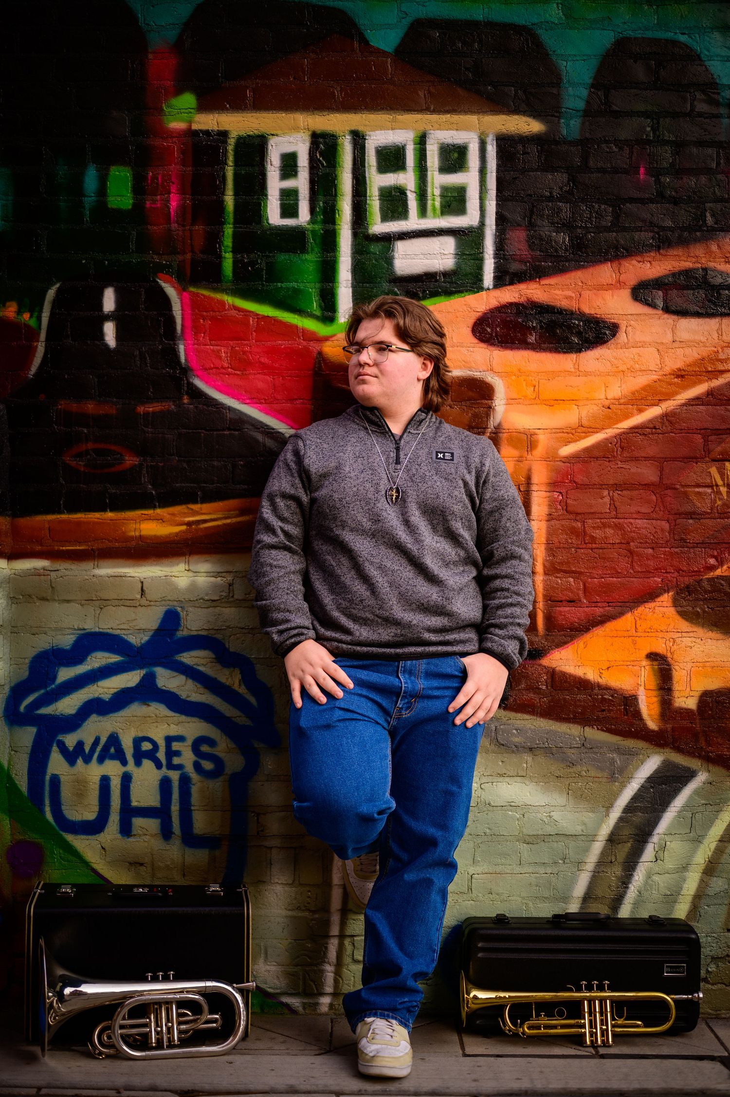 A young musician poses with trumpet cases against a colorful urban graffiti mural wall wearing a gray pullover.