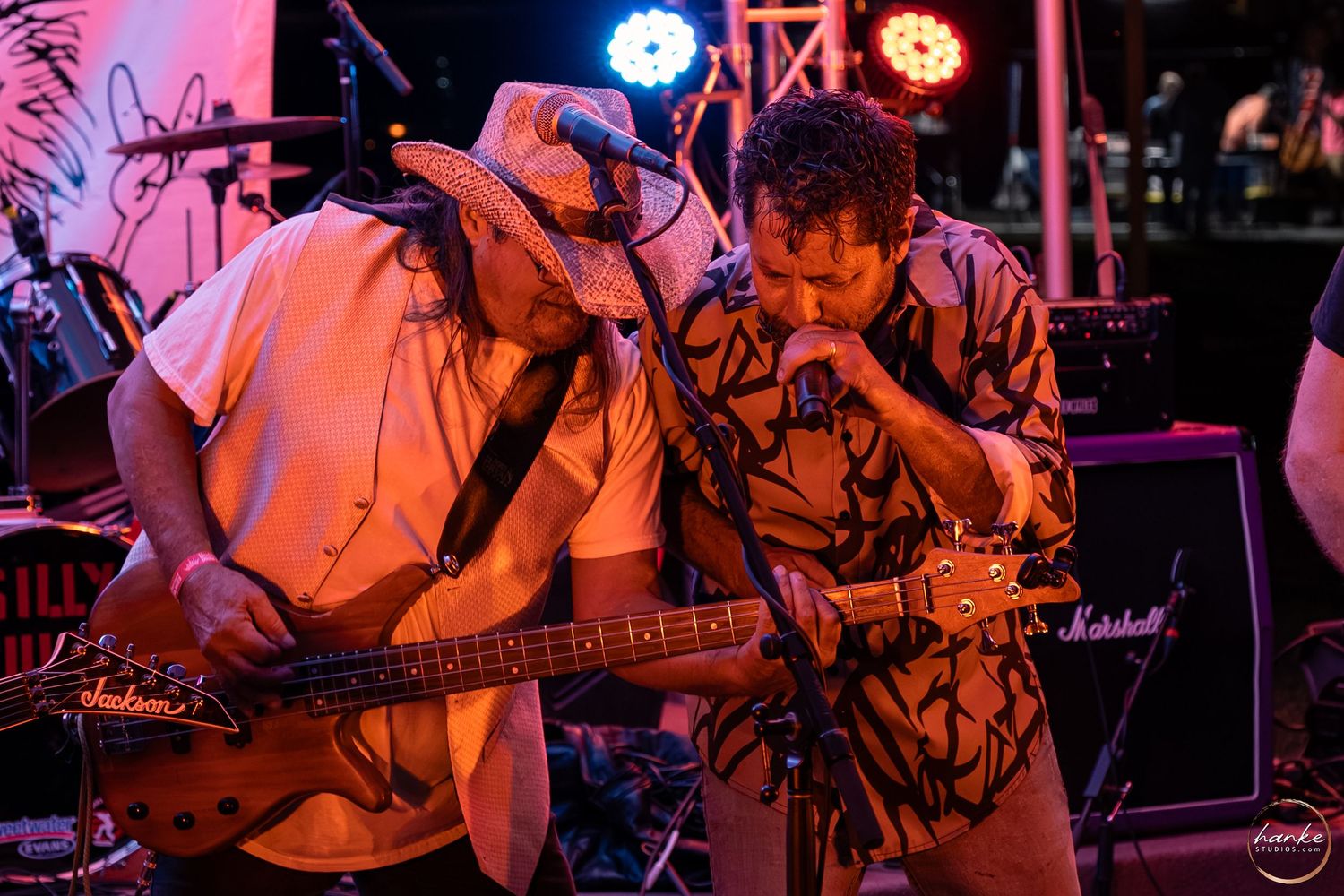 Two musicians playing guitars together on stage under warm pink lights with Marshall amp in background.