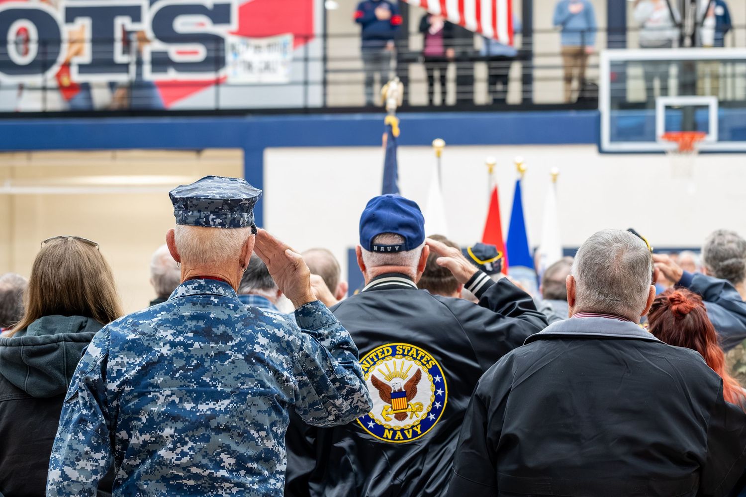 Military veterans saluting at a patriotic ceremony indoors, wearing Navy uniforms and veteran jackets with U.S. flags displayed.