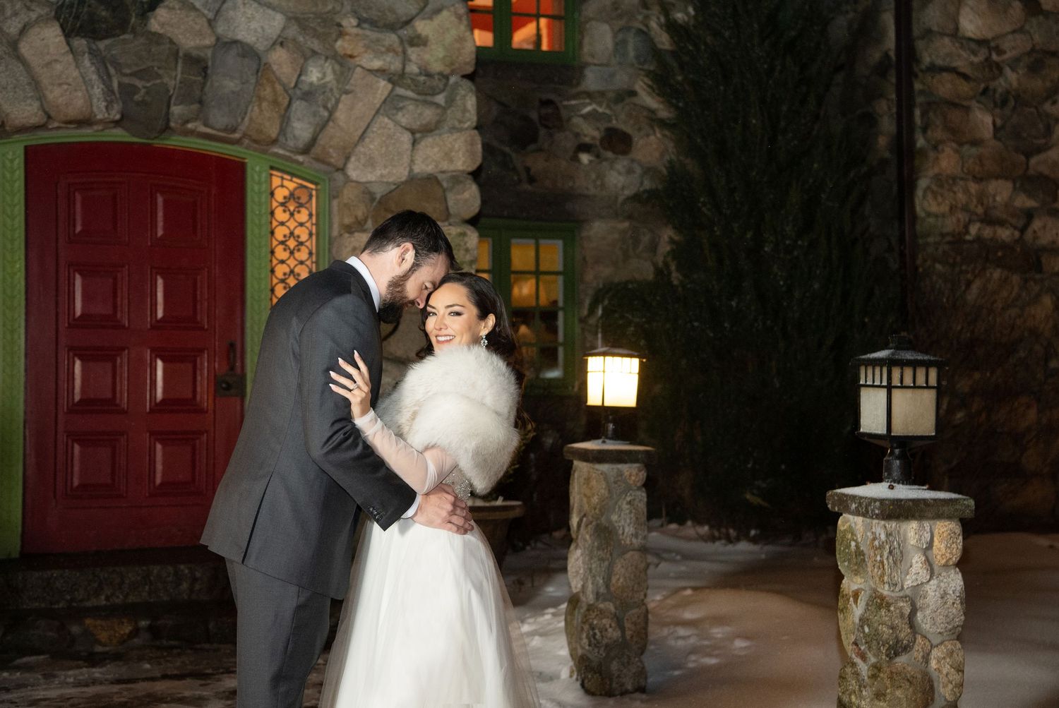 A romantic couple shares their first dance as newlyweds in front of a rustic stone wall and red door.
