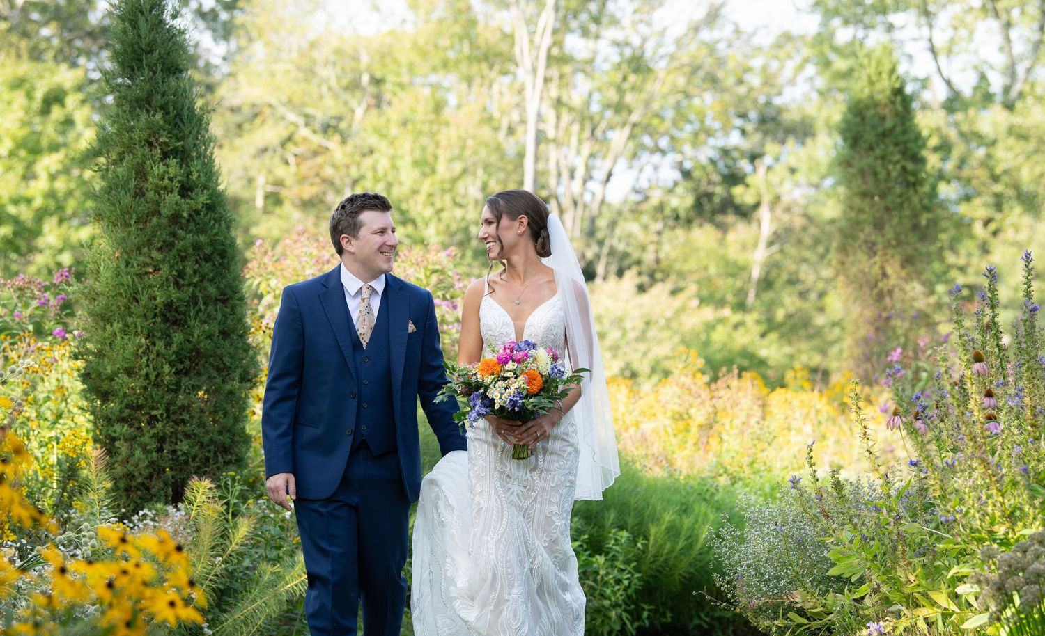 A couple in wedding attire walks through a lush garden path with evergreen trees and yellow wildflowers in bloom.