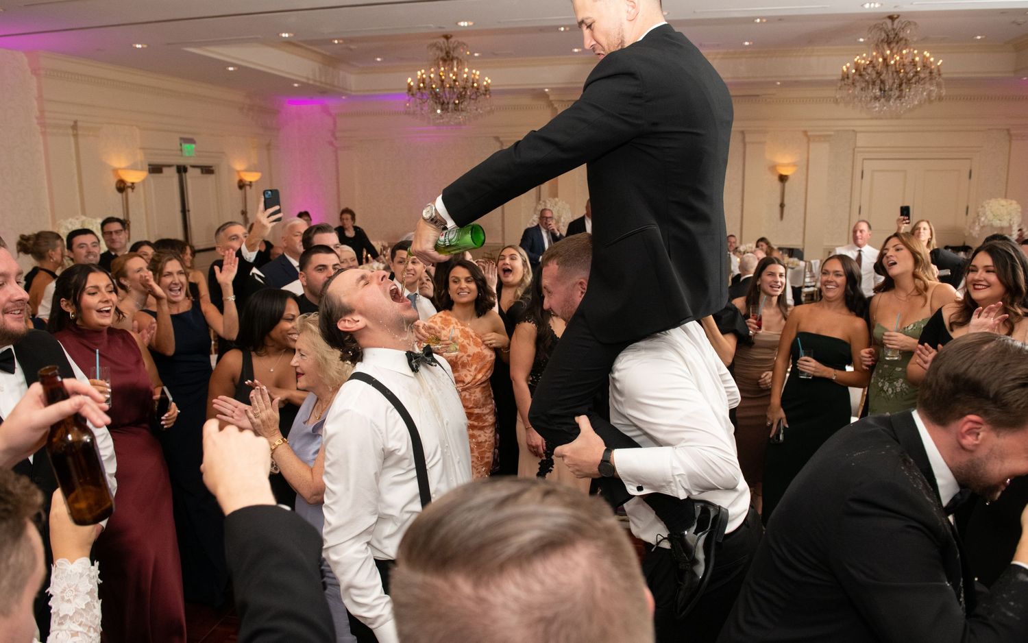 Wedding guests dance and celebrate on a crowded ballroom dance floor under elegant chandeliers at a formal reception.