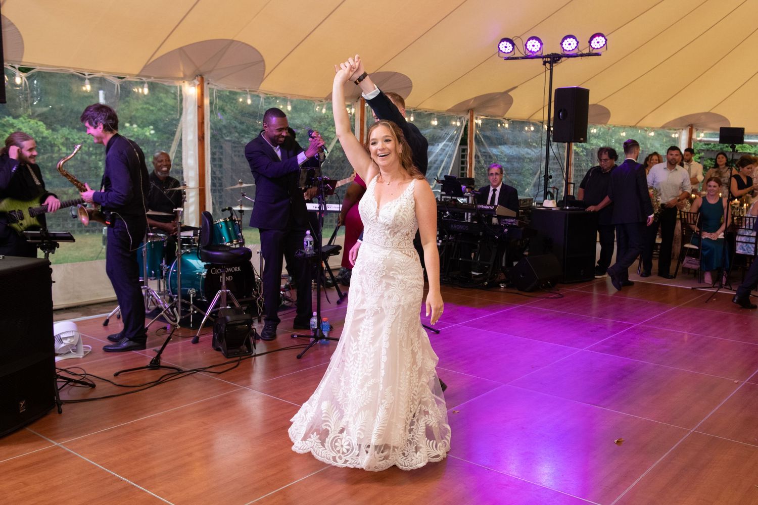 A bride dances under a tent at an evening wedding reception with purple lighting and a band in the background.