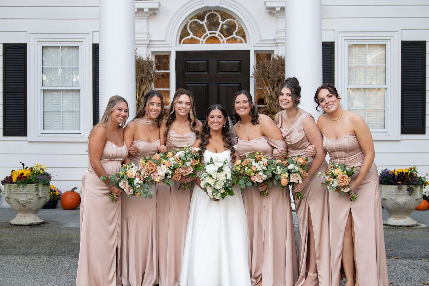 Bridesmaids in blush pink dresses stand together holding white and green floral bouquets at an outdoor wedding venue.
