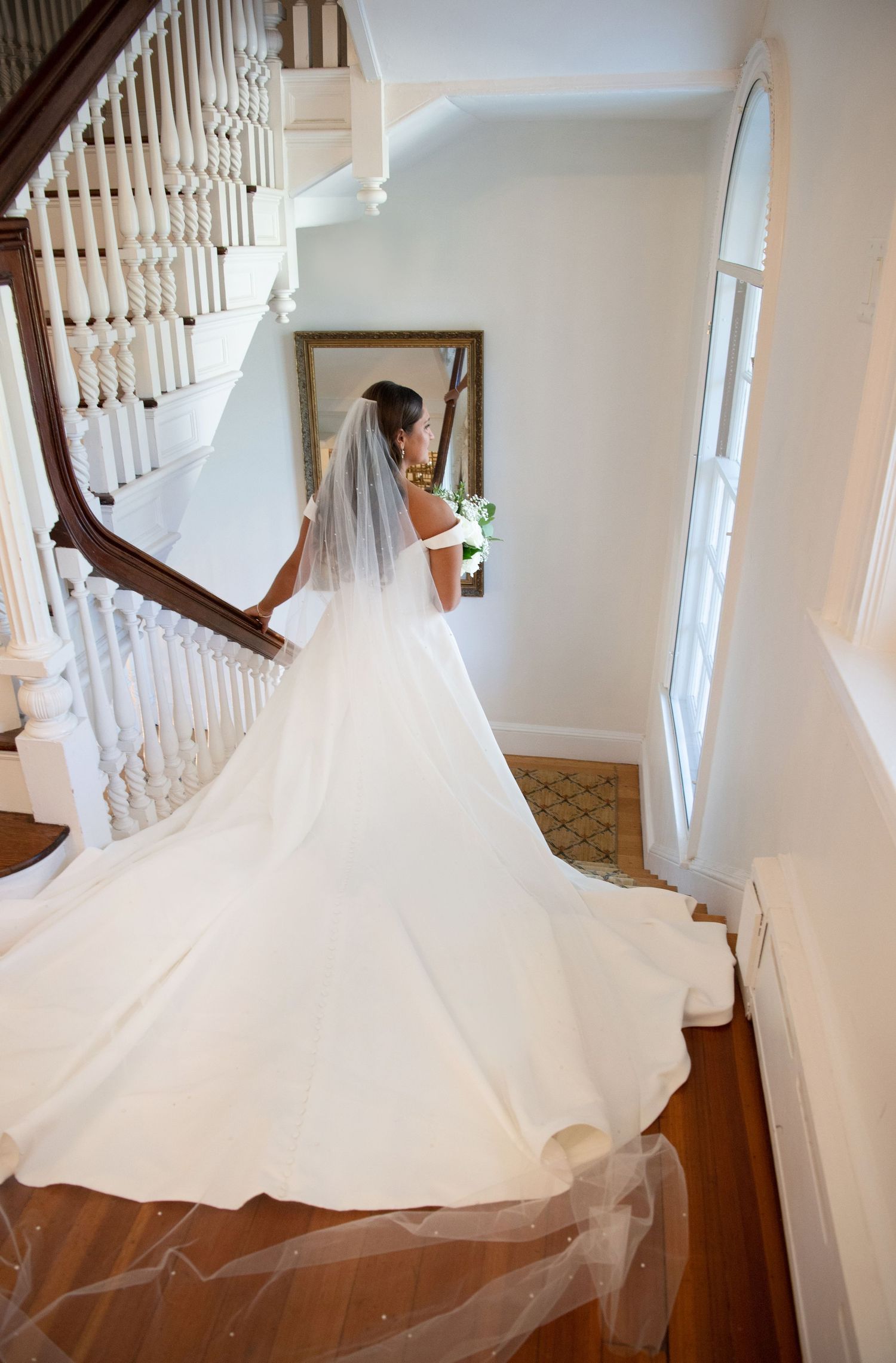 A bride in a flowing white wedding gown with a long cathedral veil stands on an elegant staircase with white railings.