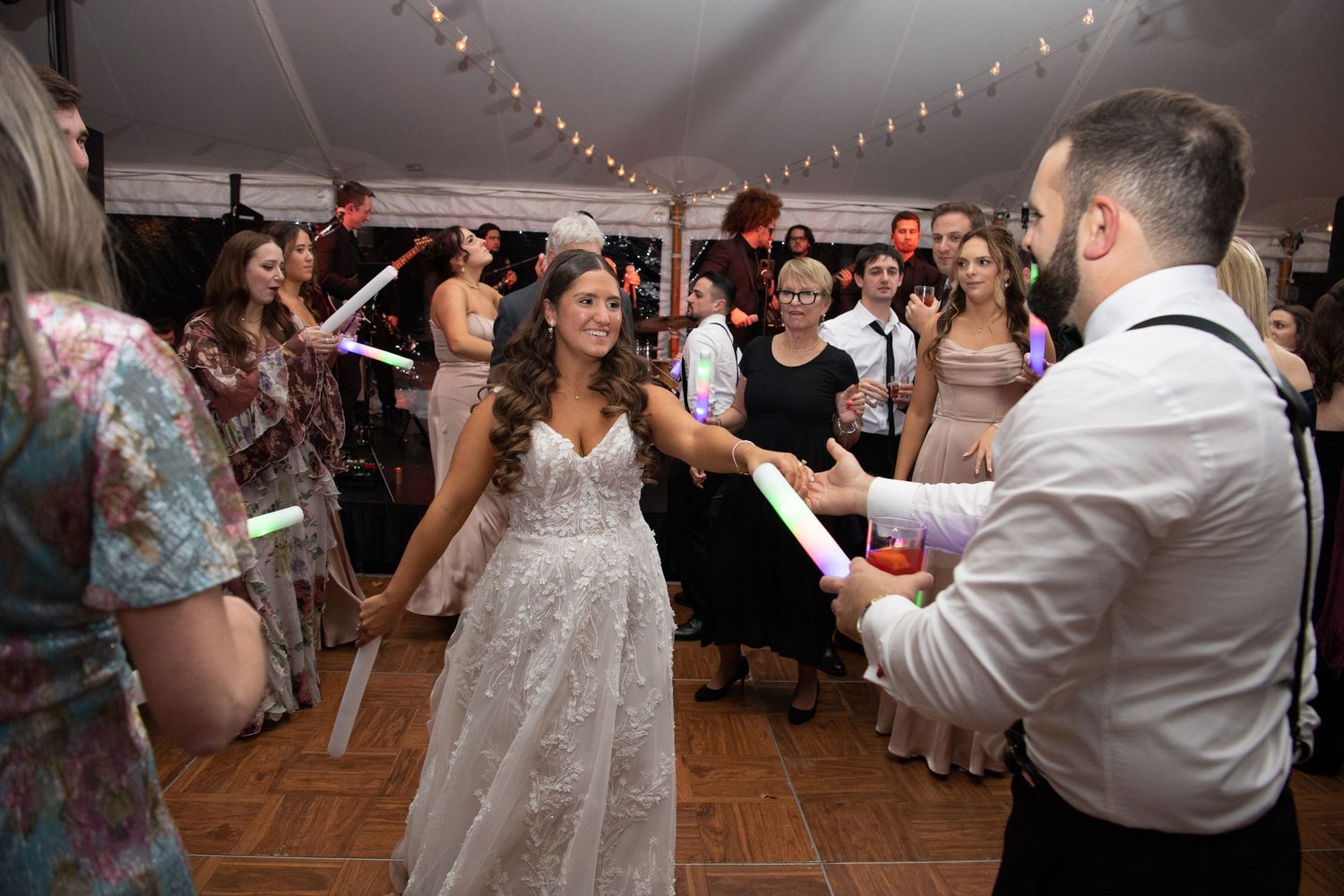 Wedding guests dance and celebrate on a wooden dance floor under string lights at an indoor reception.