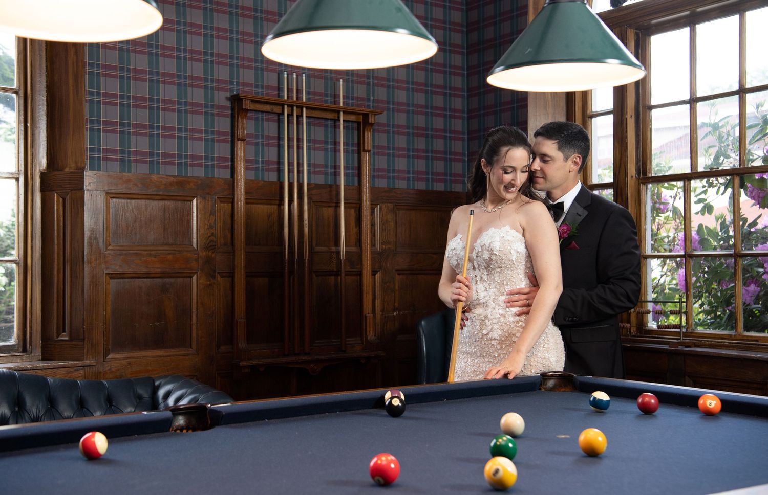 Bride and groom pose elegantly beside a billiard table in a wood-paneled room with plaid walls and pendant lights.
