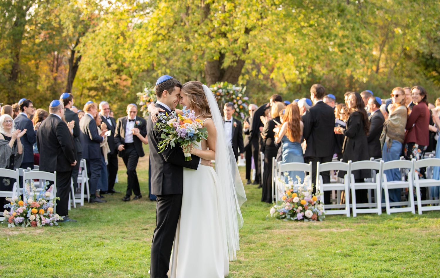 A panoramic view of an outdoor wedding ceremony in a park setting with white chairs arranged on a lawn.