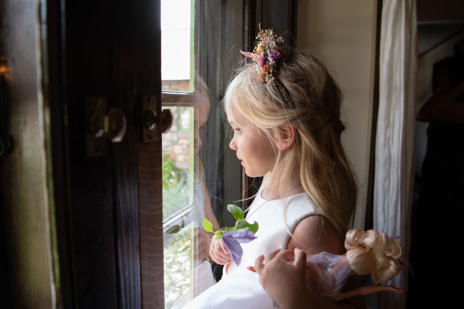 A person in a white dress gazes thoughtfully out of a window in a dimly lit room.