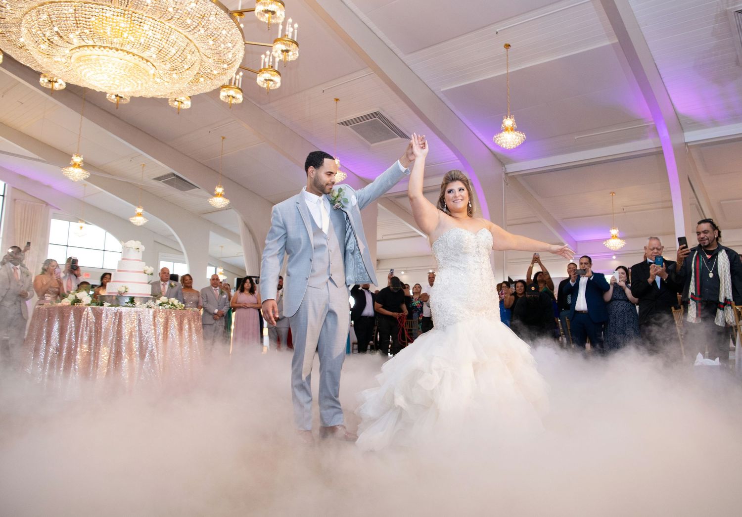 Wedding guests dance and celebrate in a grand white ballroom with elegant chandeliers and purple accent lighting.