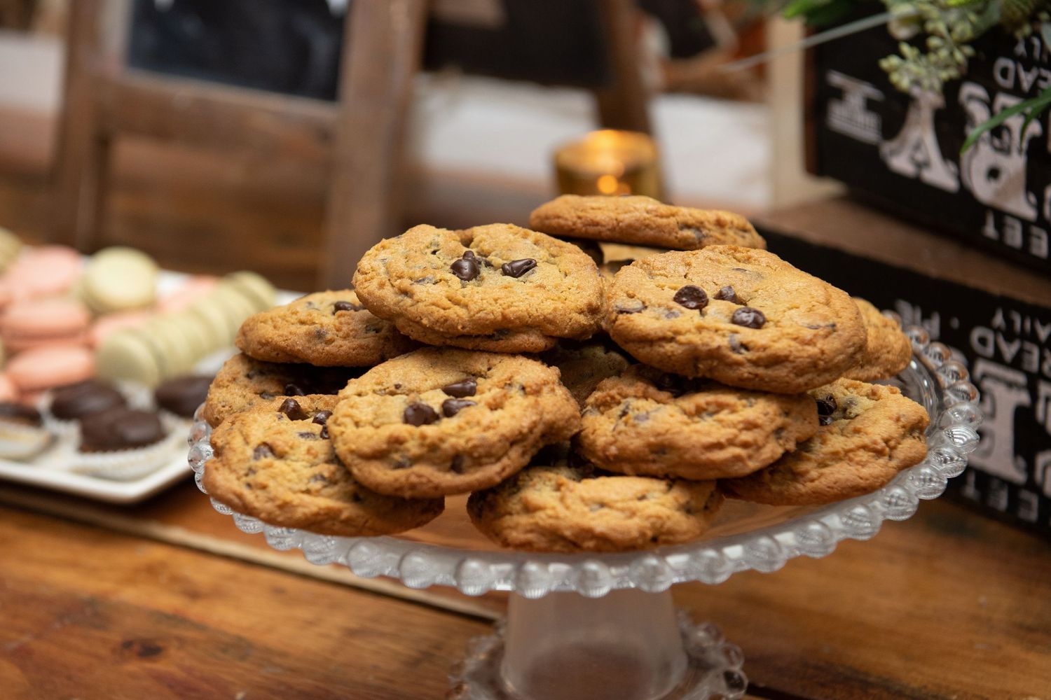Freshly baked chocolate chip cookies stacked on a glass serving plate in a rustic kitchen setting.