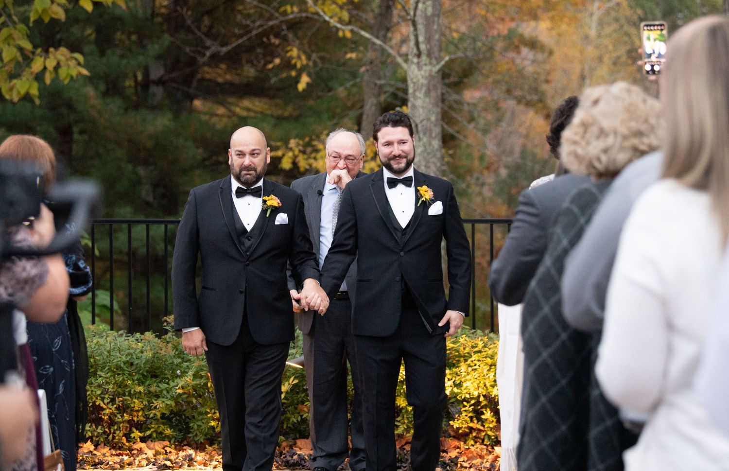 Wedding party members in black tuxedos stand in a row outdoors with autumn foliage in the background.