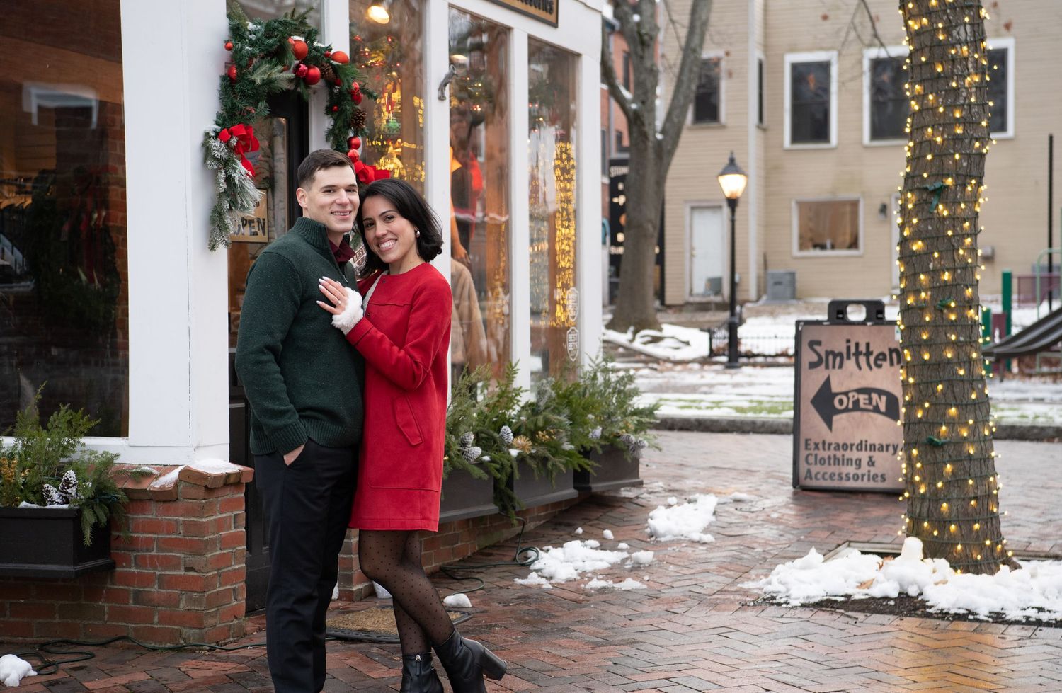 People stroll along a snow-covered brick walkway decorated with holiday lights and garland on winter evening.