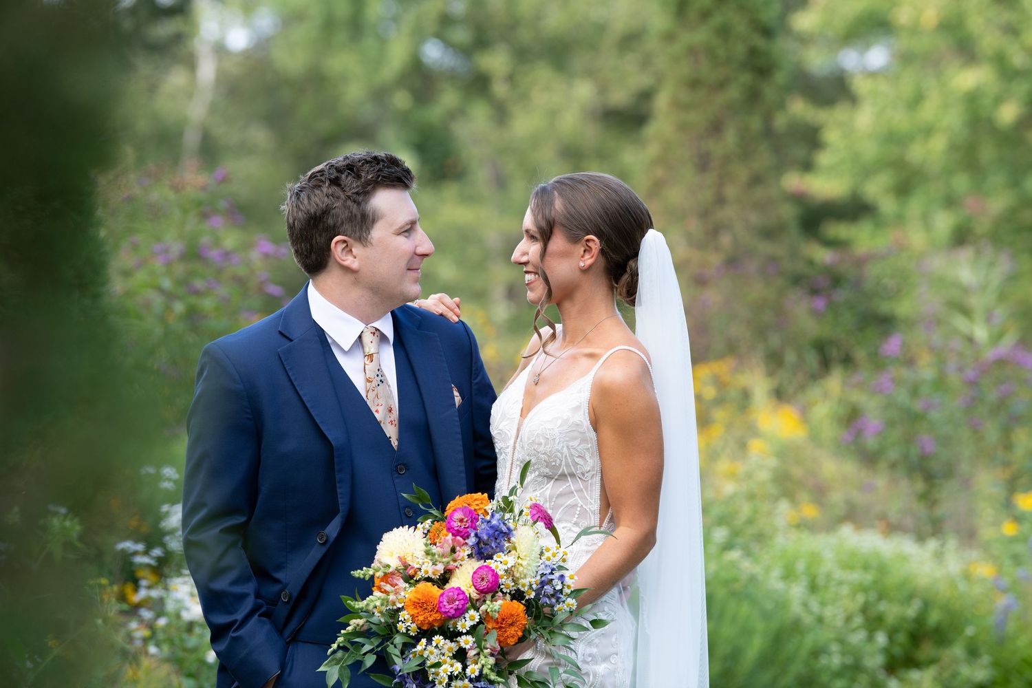 A couple in wedding attire gazes at each other while holding a vibrant bouquet in a lush garden setting.