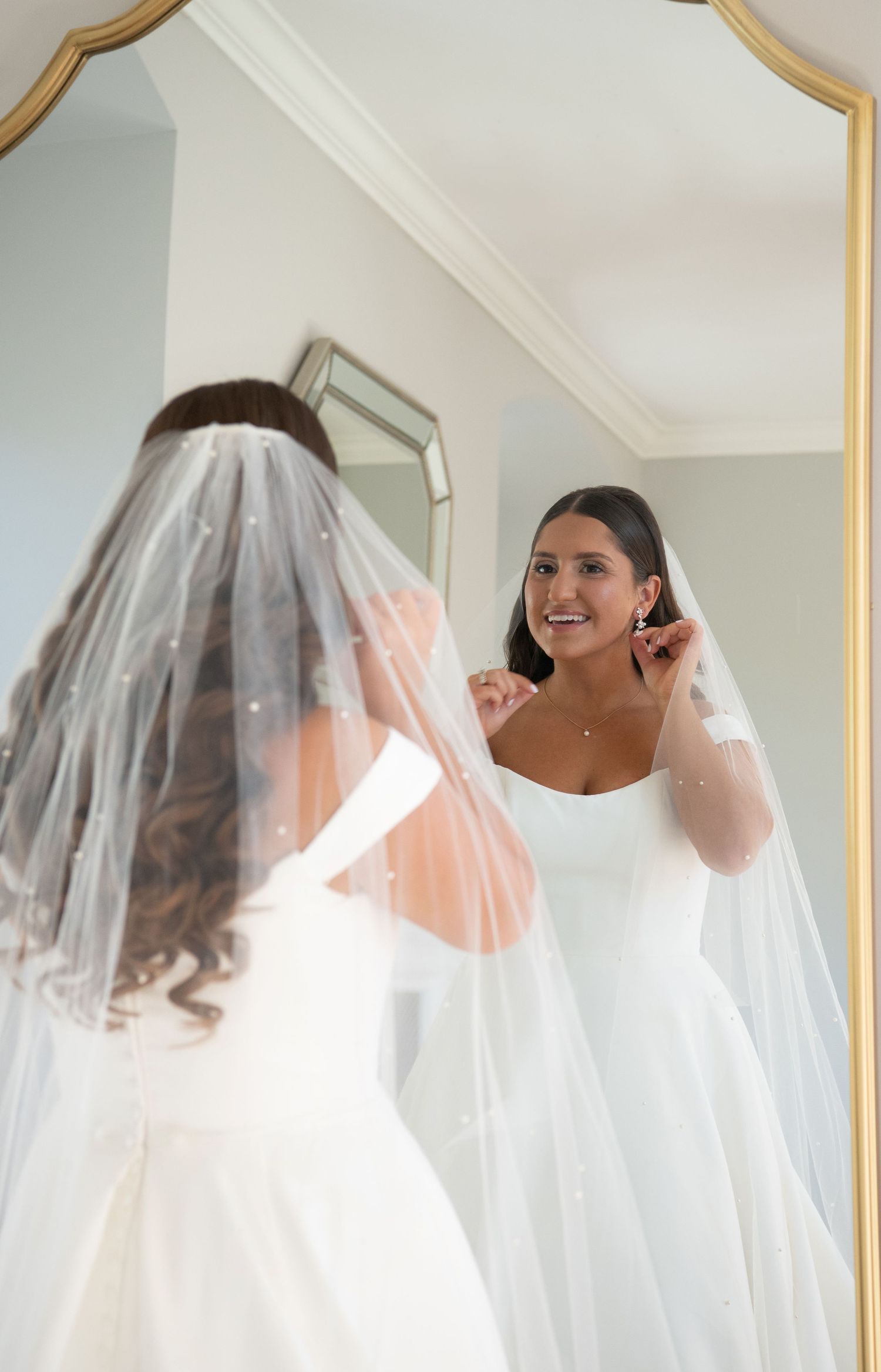 A bride in a white wedding dress gets ready for her special day, adjusting her flowing veil in a bright room.