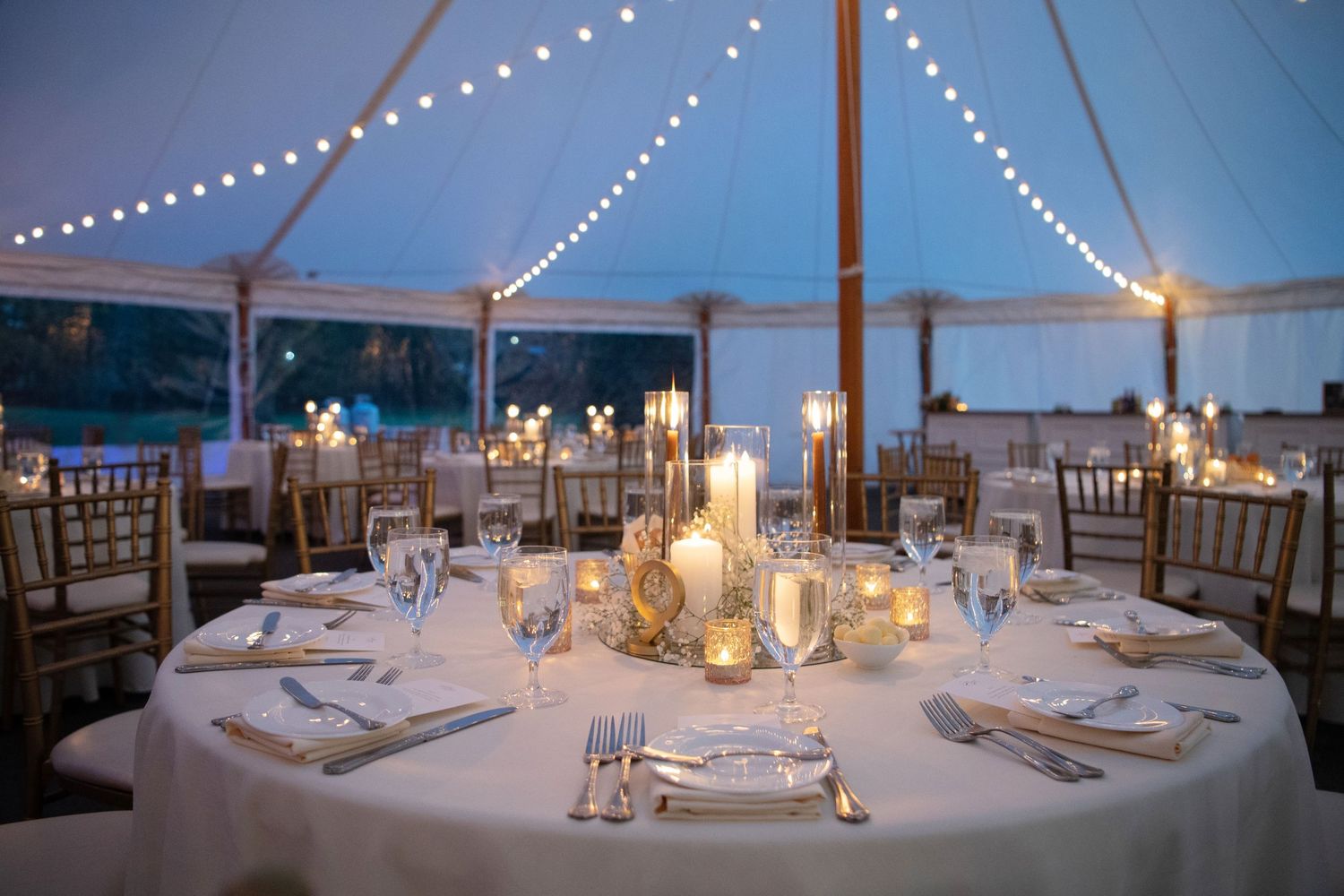 String lights illuminate an elegant outdoor wedding reception setup with white tablecloths and glowing lantern centerpieces.