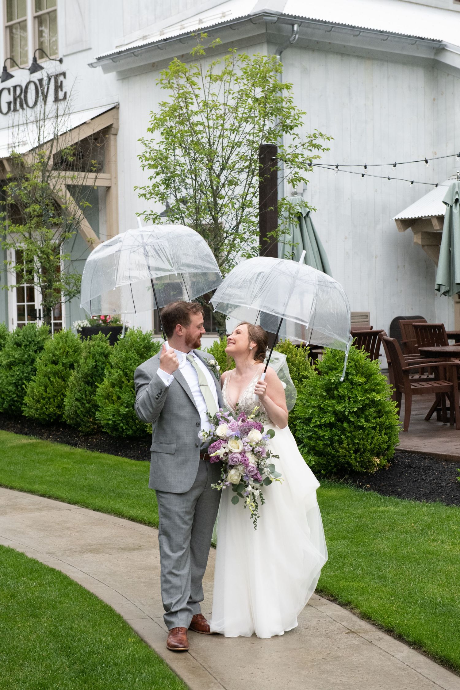A couple shares a romantic moment under a clear umbrella on a rainy wedding day outside a white building.