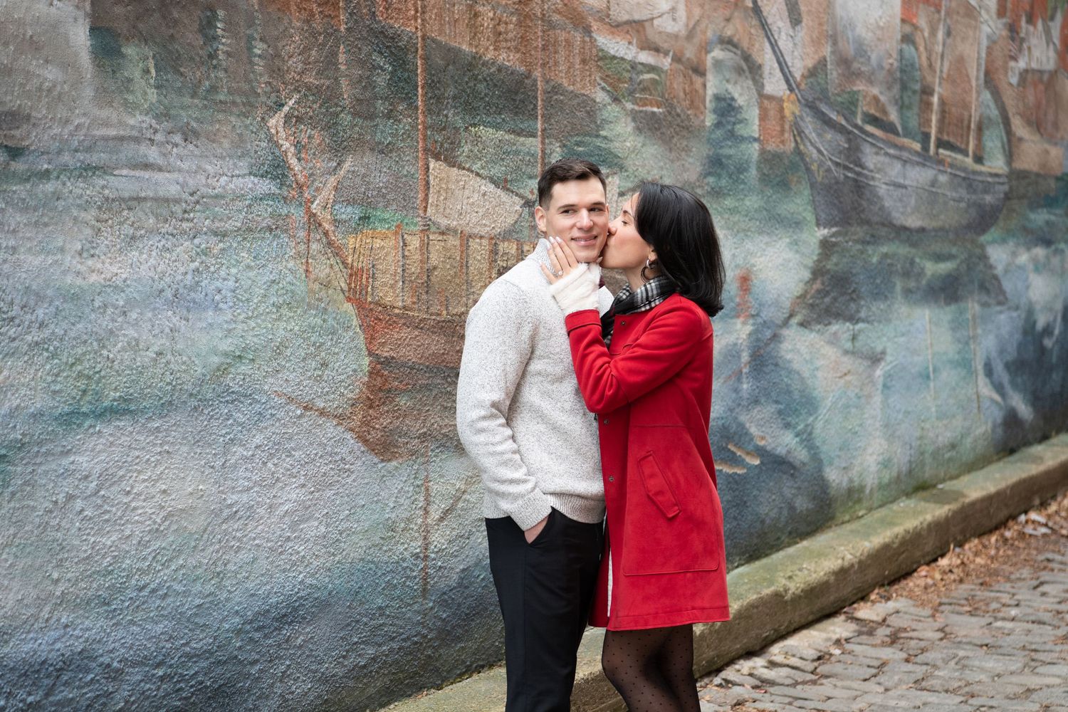 A couple shares a romantic moment against a colorful mural wall, with one wearing a red coat and the other in white.