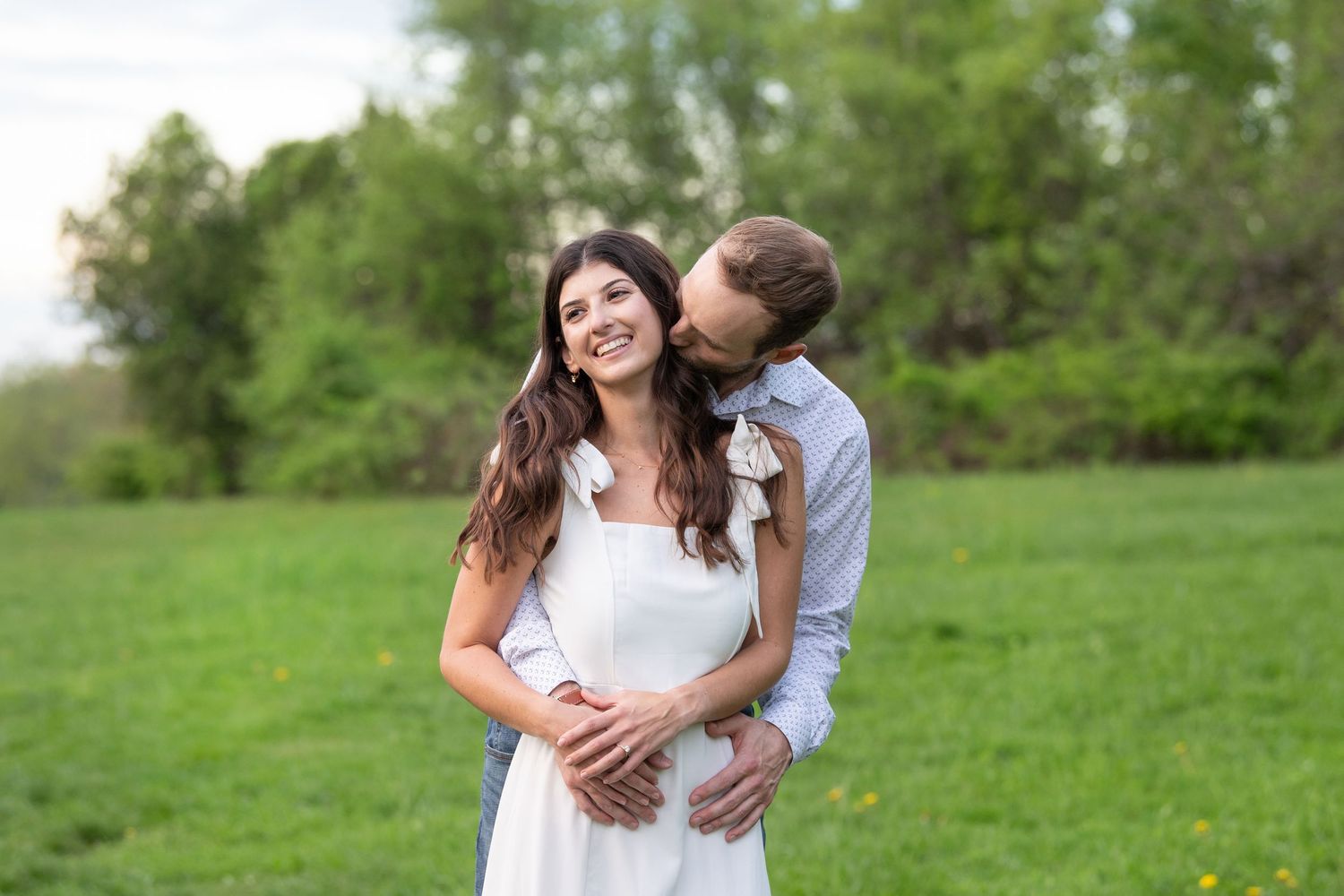 A romantic couple embraces in a sunlit meadow with green foliage in the background.