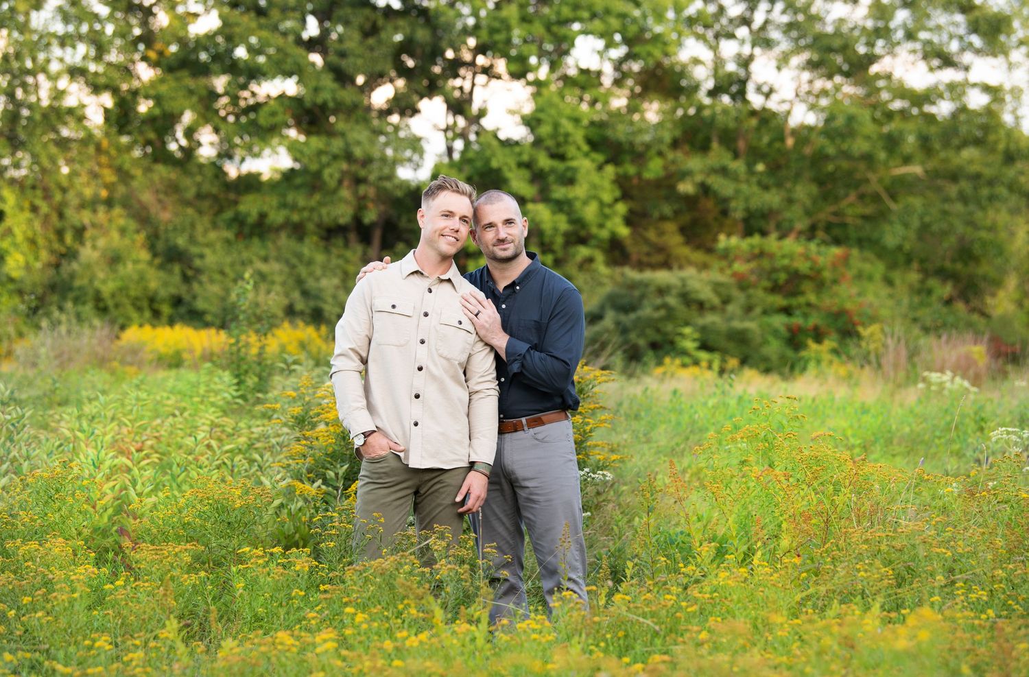 A couple stands together in a vibrant meadow filled with yellow wildflowers on a sunny summer day.