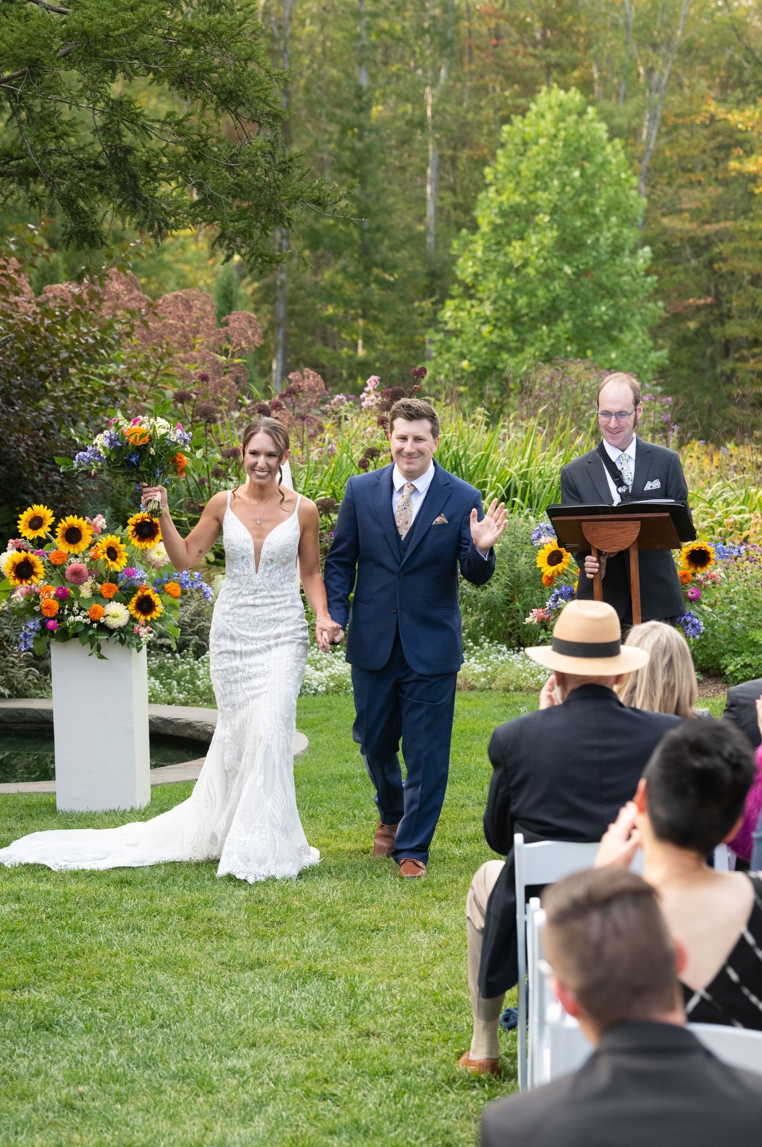 Bride and groom walk down the aisle at an outdoor garden wedding ceremony, smiling and waving at guests.