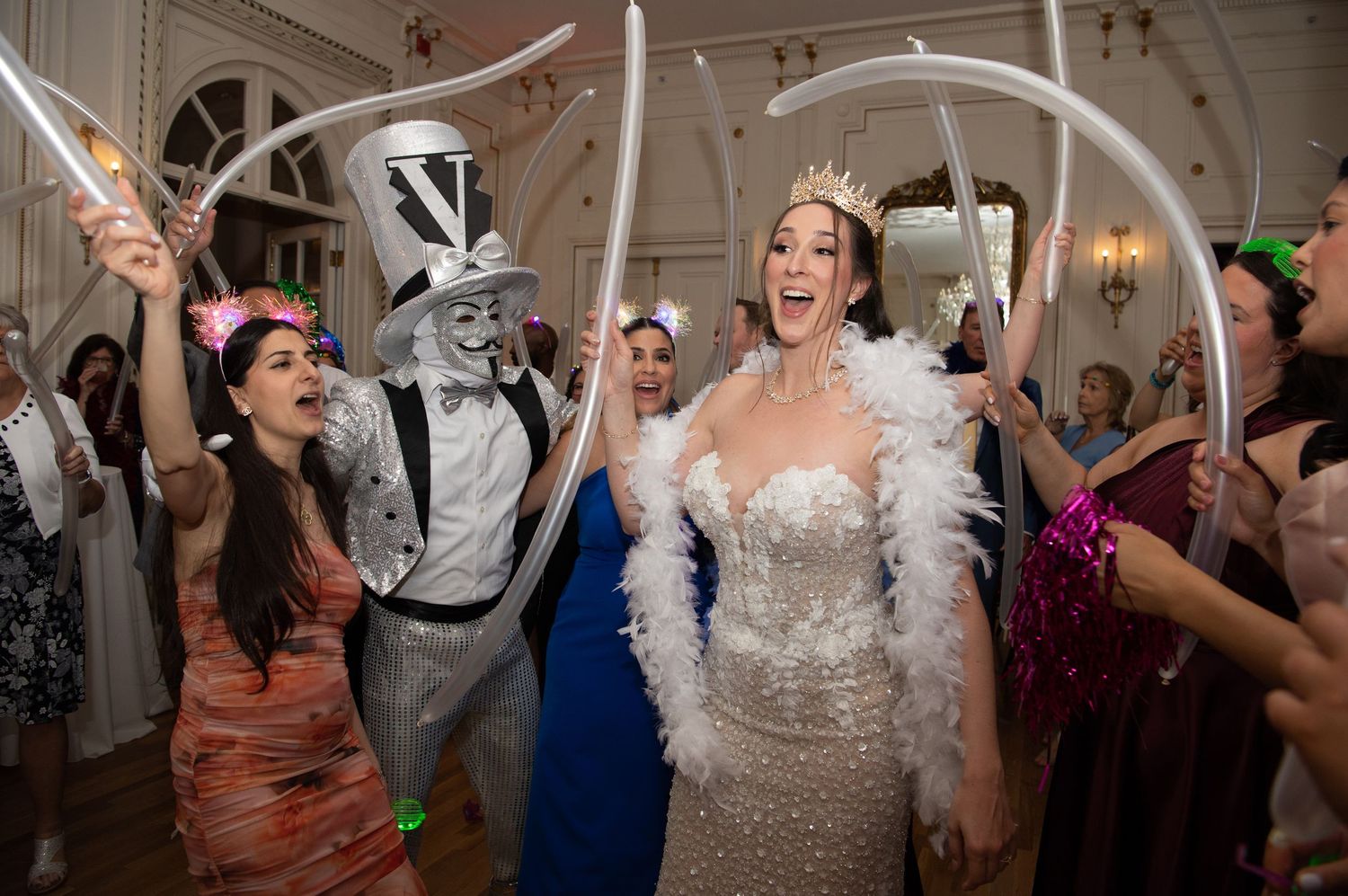 Bride in sparkling gown and crown celebrates with guests holding balloon arches at lively wedding reception.