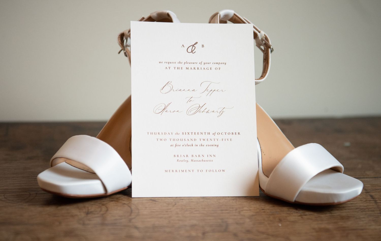 Elegant white wedding shoes arranged with delicate invitation cards and floral details on a wooden surface.