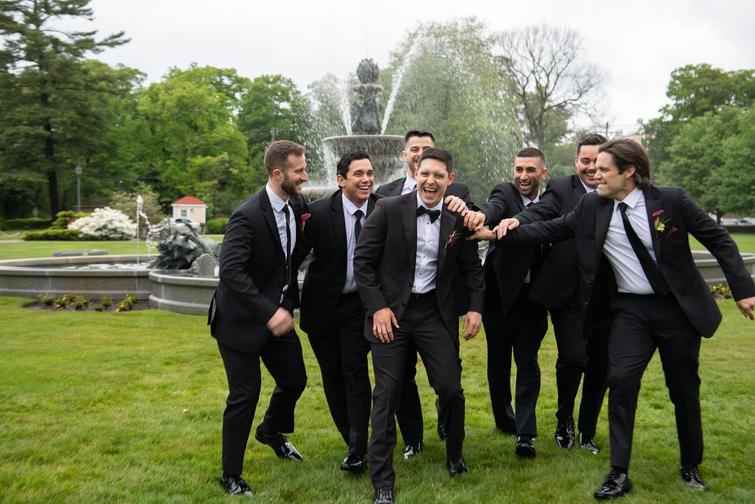 Groomsmen in black tuxedos laughing and celebrating near a fountain in a lush green park on a wedding day.