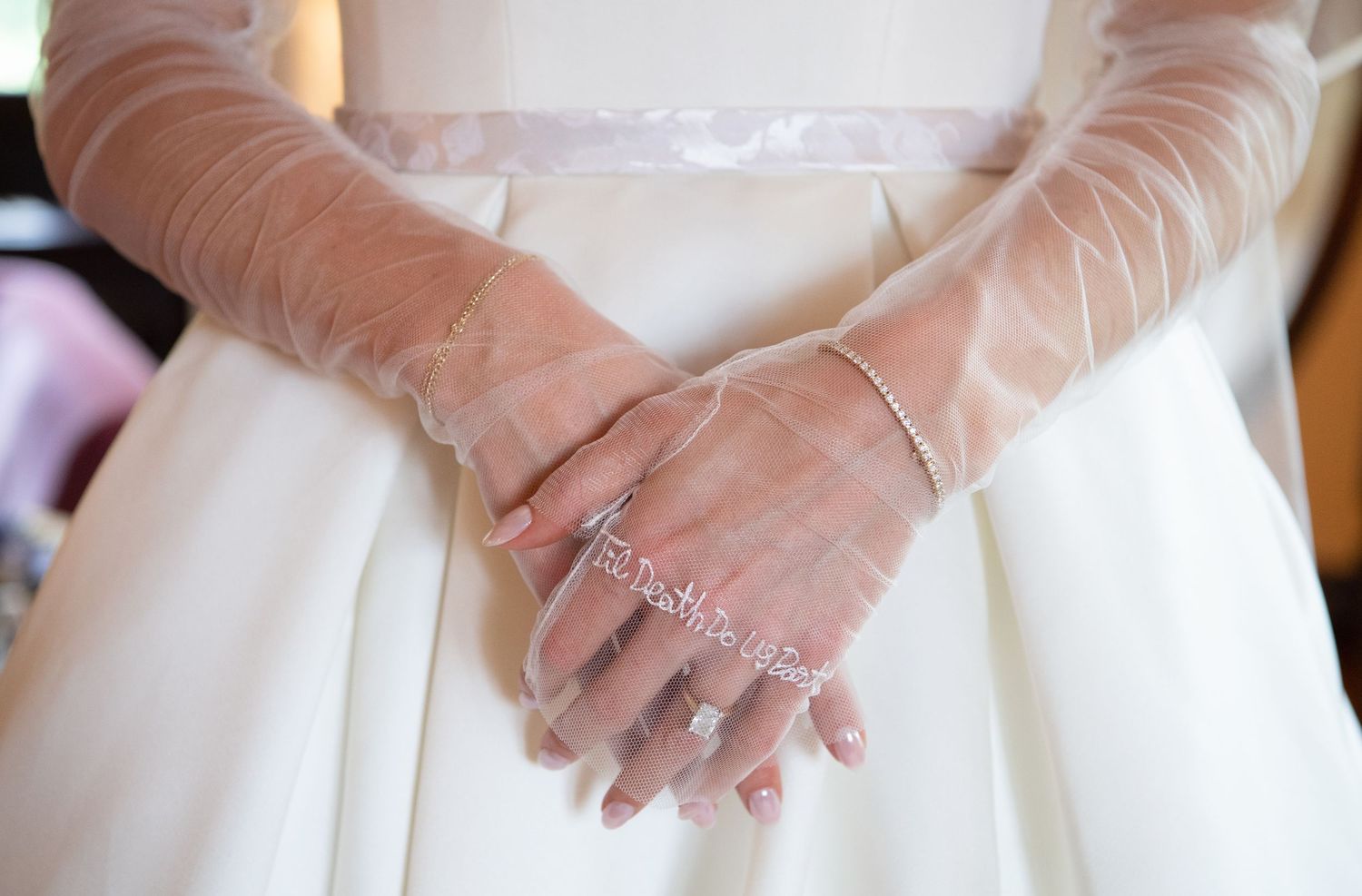 Bride wearing sheer lace gloves and a delicate bracelet, hands clasped over white wedding dress.