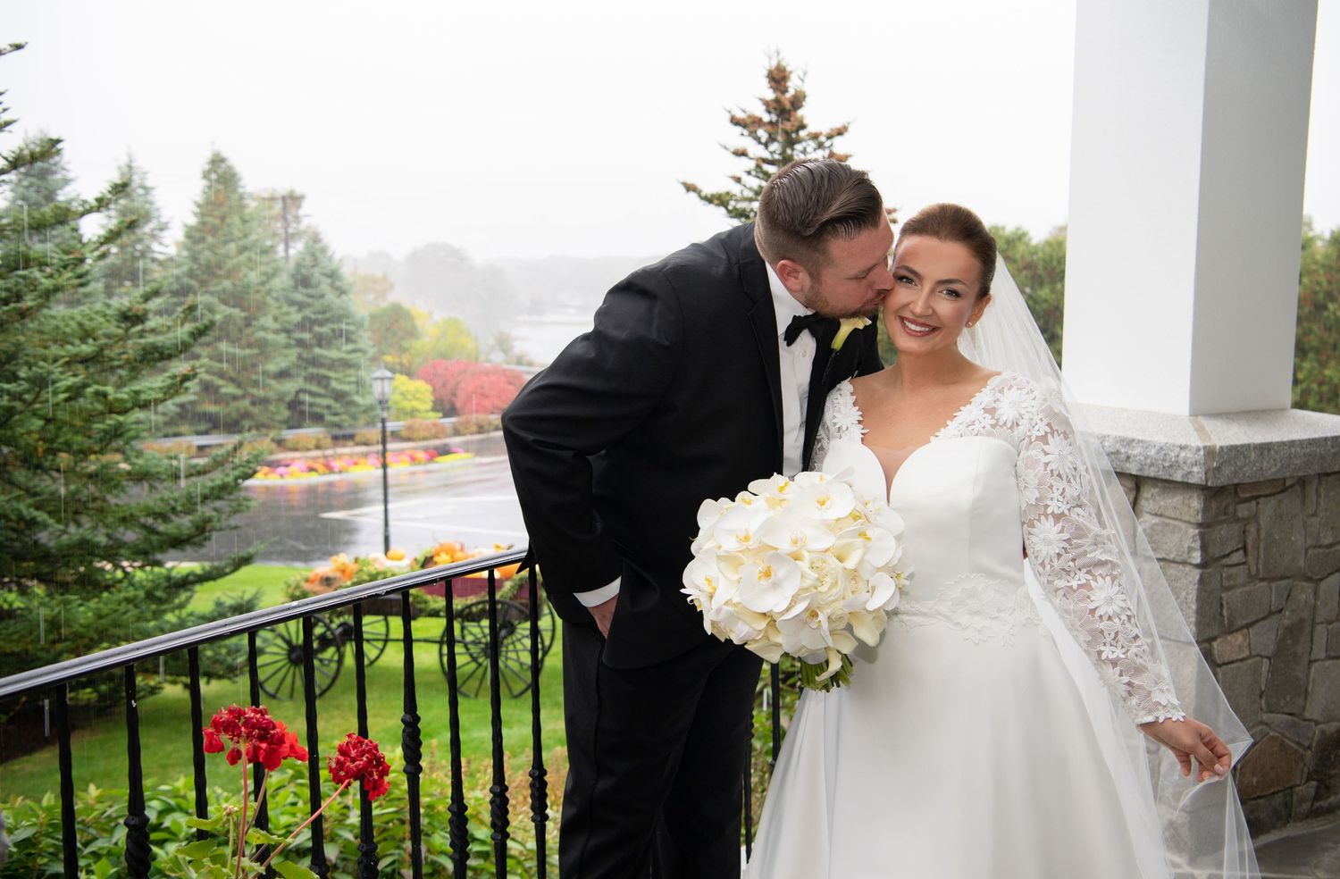 A wedding couple shares a tender moment on a porch with white columns and an autumn backdrop.