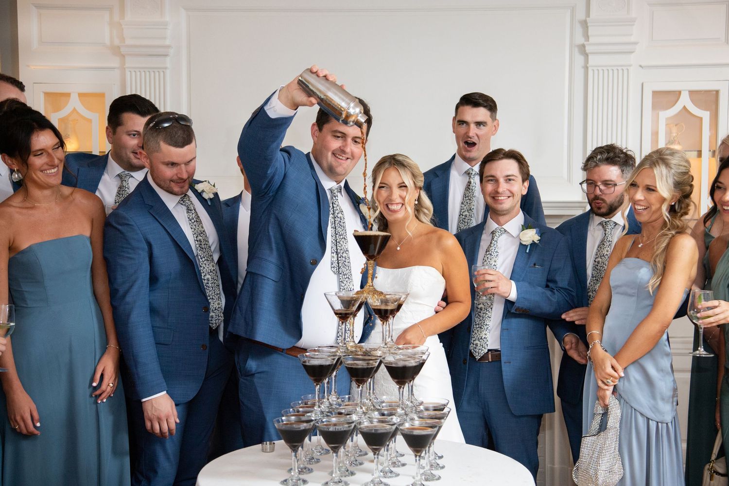 Wedding party in blue attire gathers around a champagne tower during an elegant celebration.
