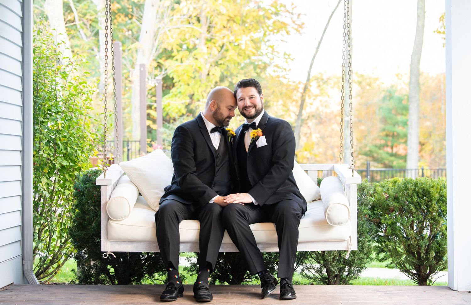 A couple in black formal attire share a romantic moment while sitting on a white porch swing surrounded by autumn foliage.