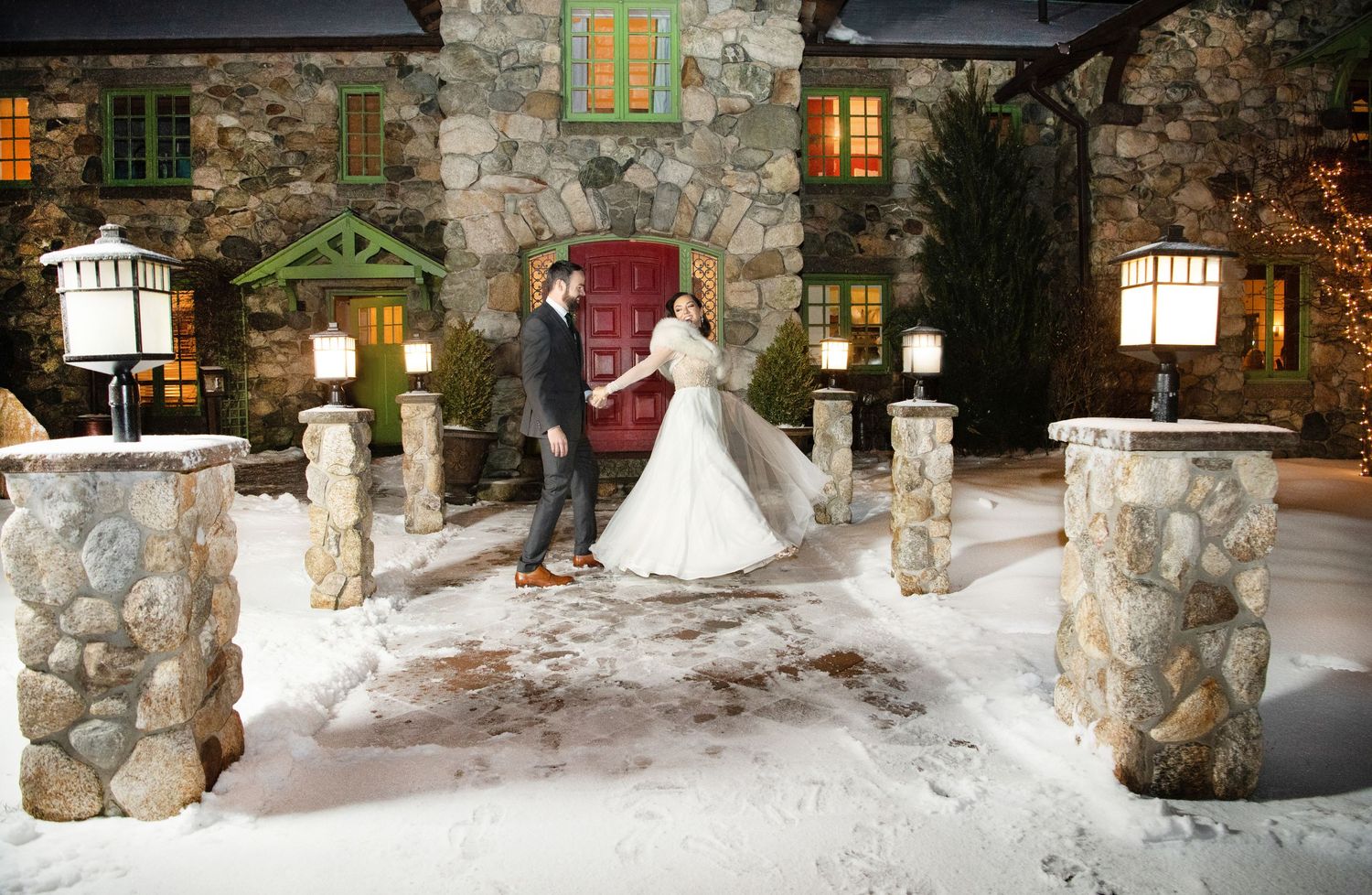 A snowy winter scene shows two figures on a porch lined with rustic stone pillars and glowing lanterns at night.